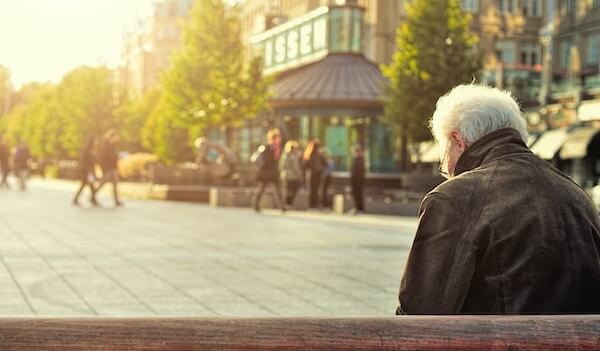 Grey haired man sits on a park bench watching others walk on a promenade. He looks lonely.